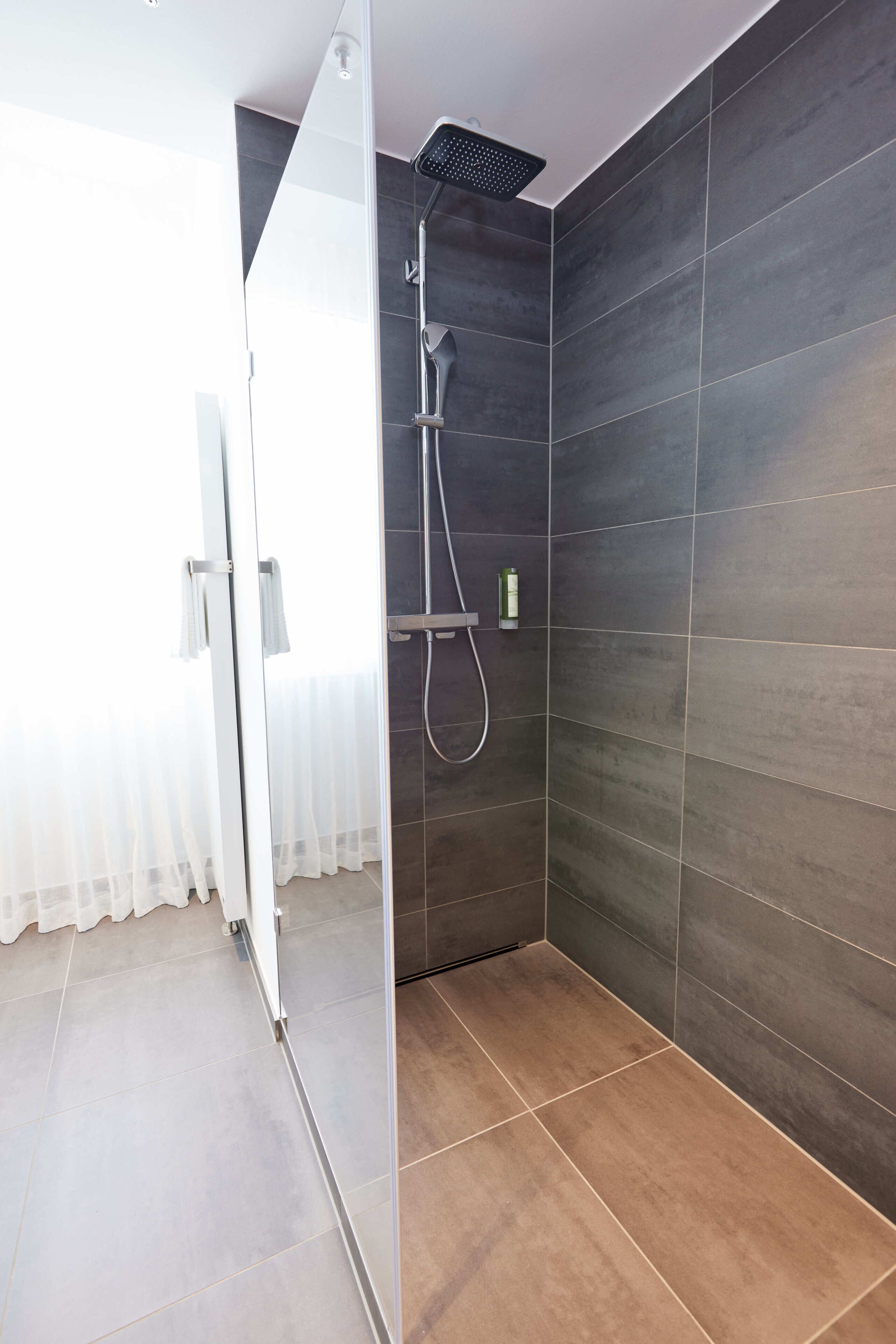 Un bagno moderno con pareti rivestite di piastrelle scure, box doccia in vetro e soffione a vista.