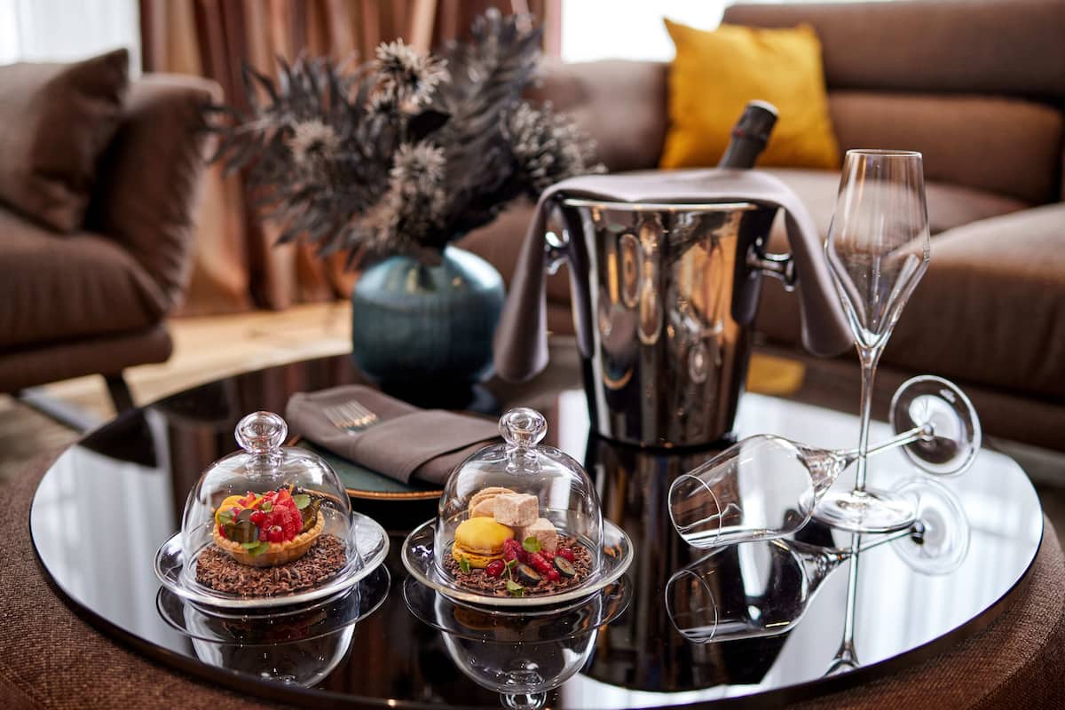 A dining table with two dessert dishes under glass domes, a silver teapot, and a wine glass.