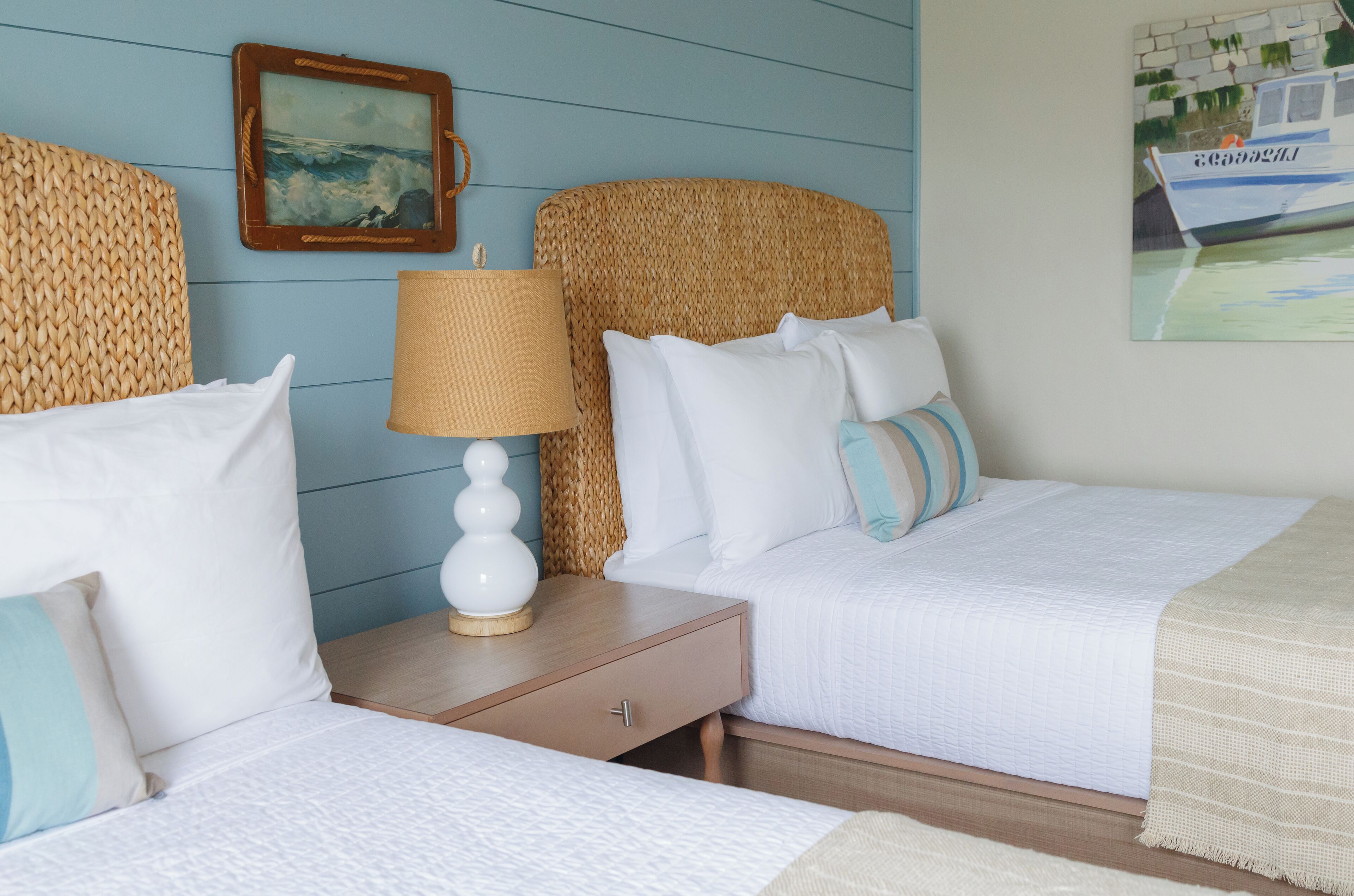 A room with two beds, a wooden nightstand with a lamp, and a woven headboard.