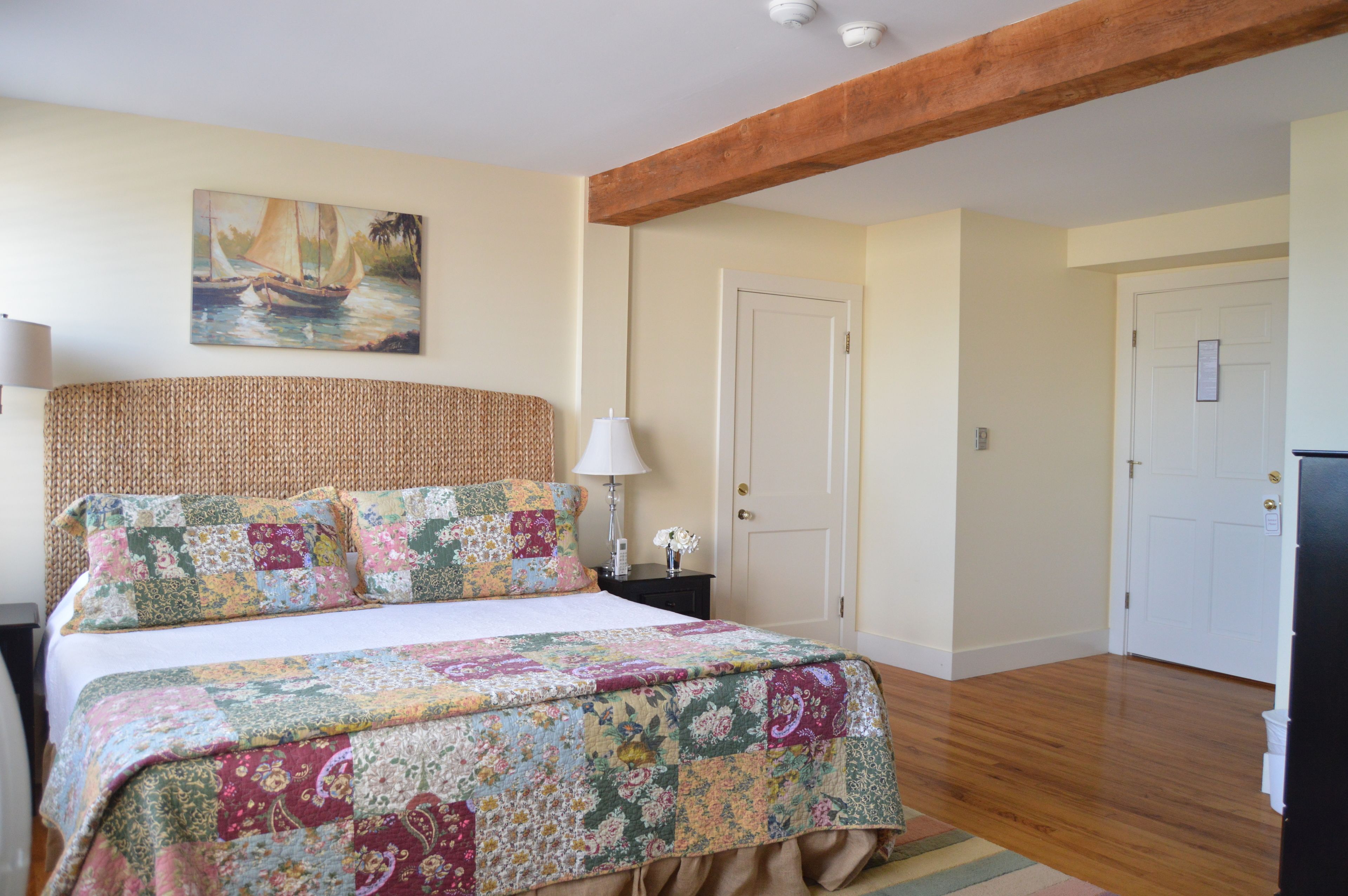A bedroom with a large bed, a wooden ceiling beam, a painting on the wall, and a door in the background.