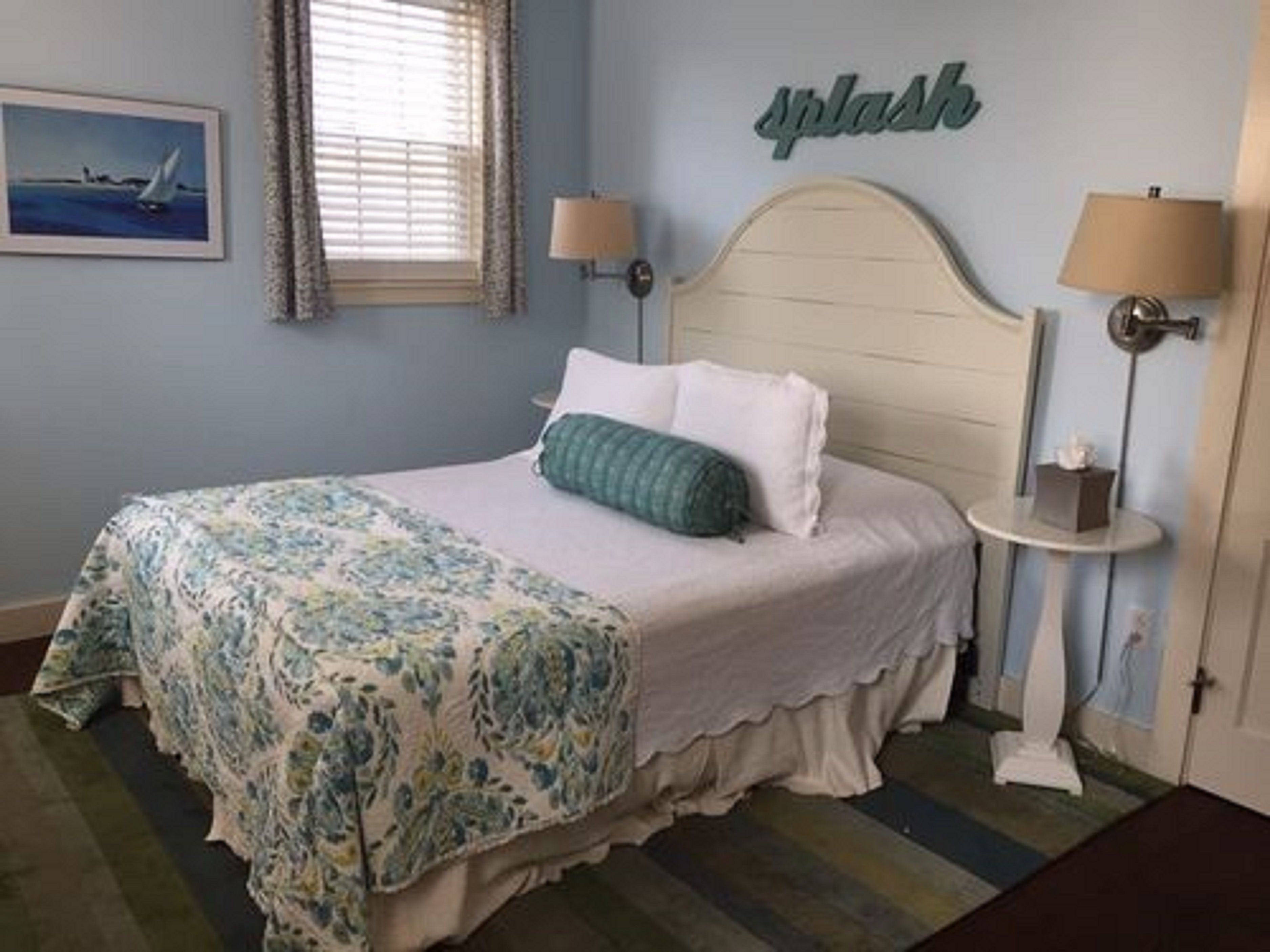 A bedroom with a bed, two bedside lamps, a window with blinds, a framed picture on the wall, and a 'Splash' sign.