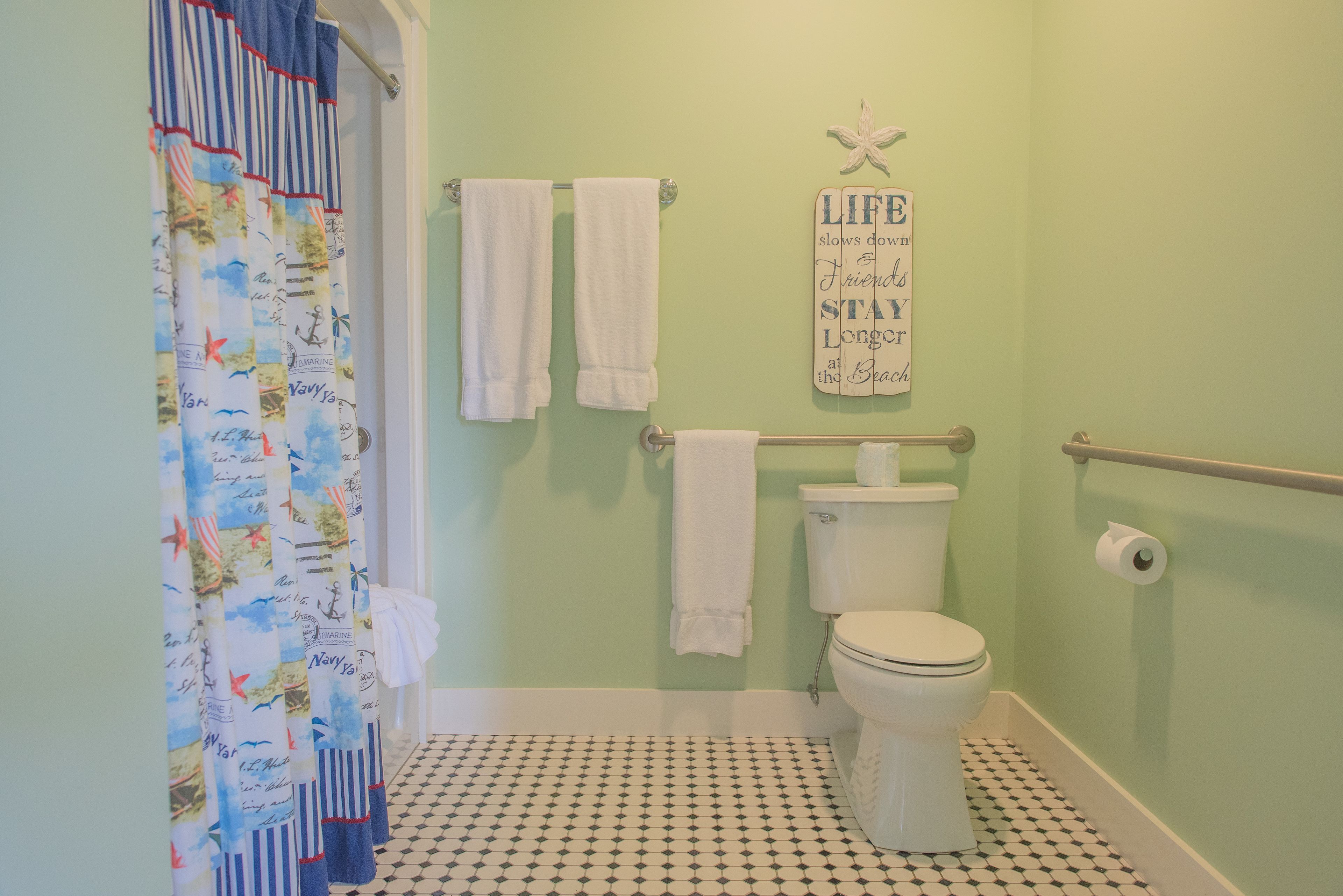 A bathroom with a nautical-themed shower curtain, a toilet, a handrail, and a motivational quote on the wall.