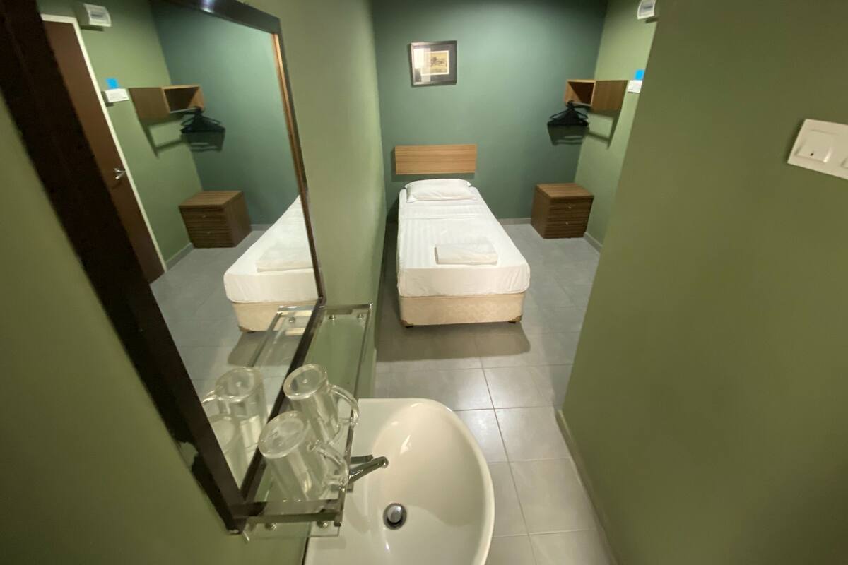 Ein Hotelzimmer mit zwei Einzelbetten, einem Badezimmer mit Waschbecken und Spiegel sowie einem kleinen Nachttisch mit Lampe.