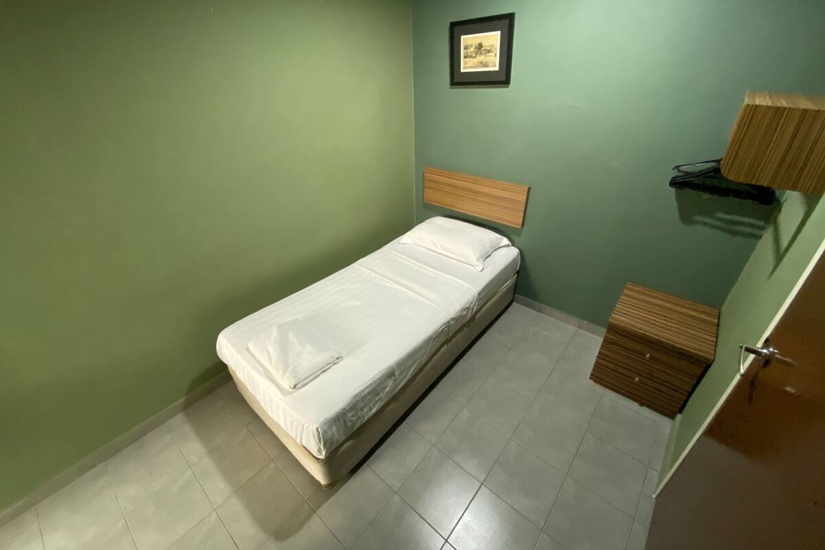 Ein kleines Zimmer mit einem Einzelbett, einem Holzkopfteil, einem gerahmten Bild an der Wand und einer kleinen Holzschublade.