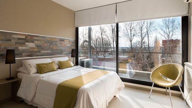 Une chambre d’hôtel moderne avec un grand lit, une vue sur la ville grâce à une grande fenêtre et un fauteuil contemporain.