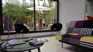 Une pièce avec une grande fenêtre, un canapé noir orné de coussins à motifs, une table ronde sur laquelle sont posés des livres et une lampe, et un fauteuil.