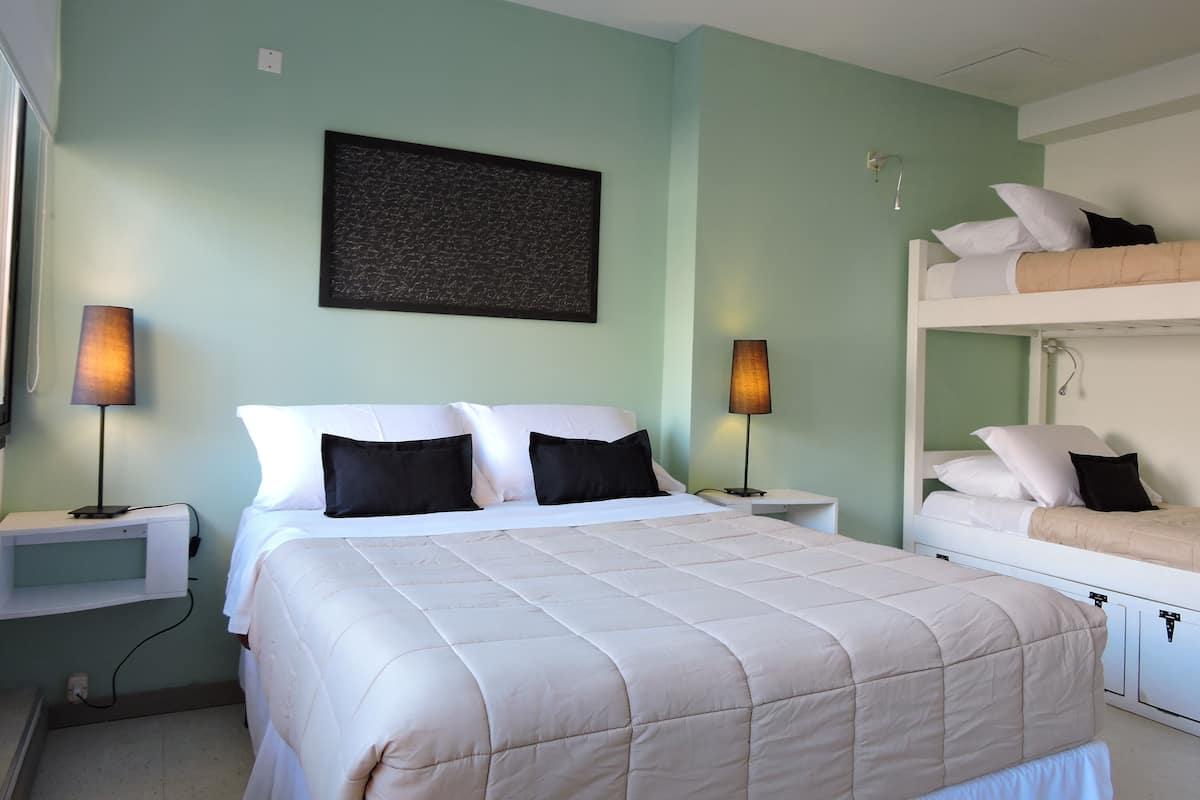 A bedroom with a bed, two bedside tables, a nightstand, a wall-mounted lamp, a bunk bed, and a framed picture on the wall.