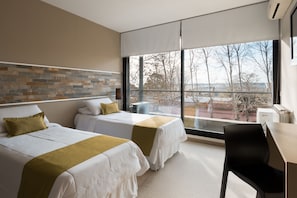 A modern hotel room with two beds, a desk, and a large window offering a view of trees and buildings.
