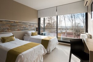 Una habitación de hotel moderna con dos camas, un escritorio y un amplio ventanal con vistas a árboles y edificios.