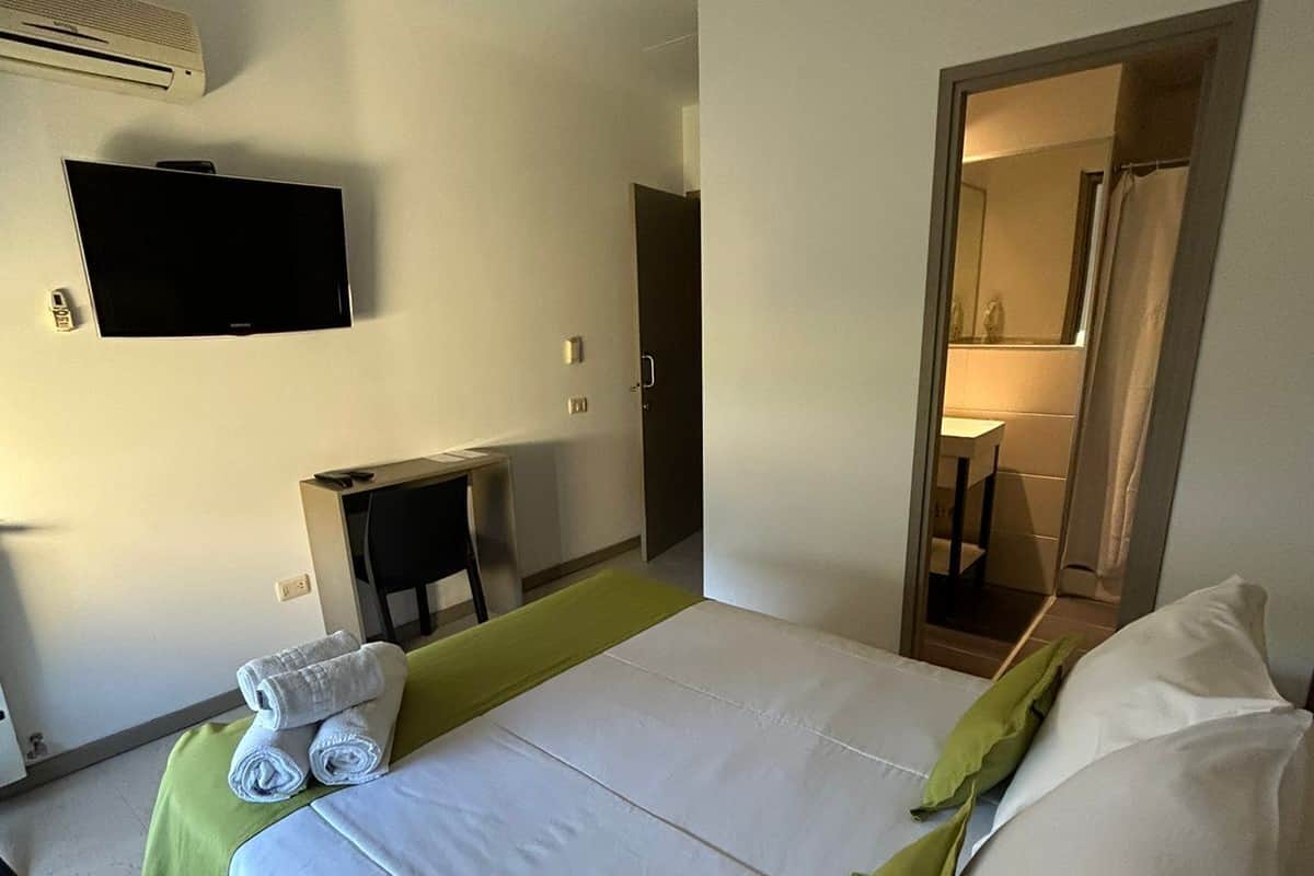 A hotel room with a bed, a flat-screen TV mounted on the wall, a desk, and a bathroom visible through an open door.
