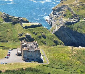 A large, historic building with a prominent entrance, situated on a lush green hillside with a view of the ocean. - Camelot Castle Hotel (Tintagel)