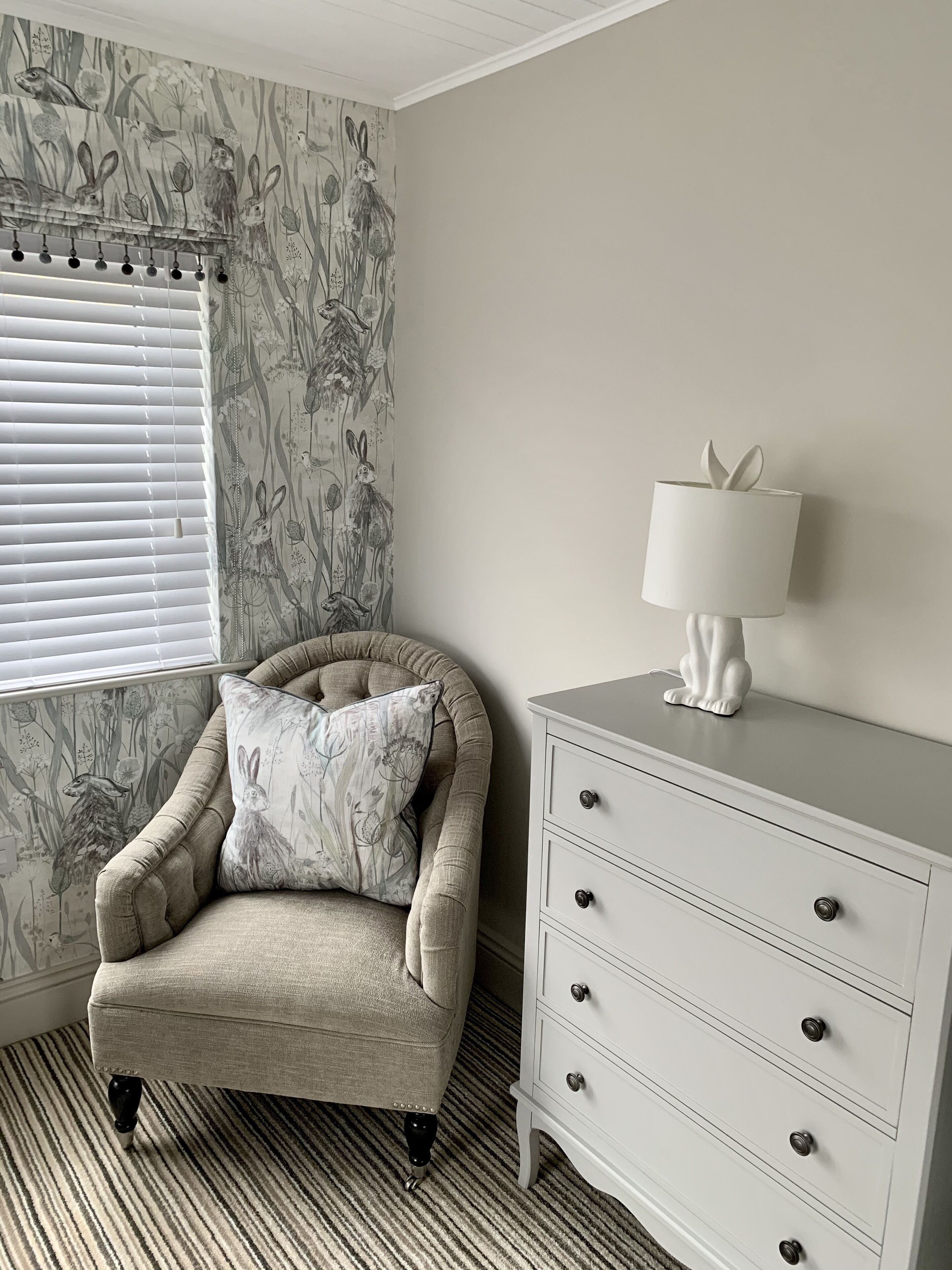 Een kamer met behang met een patroon, een beige fauteuil met kussen, een witte dressoir met lades, en een raam met jaloezieën.