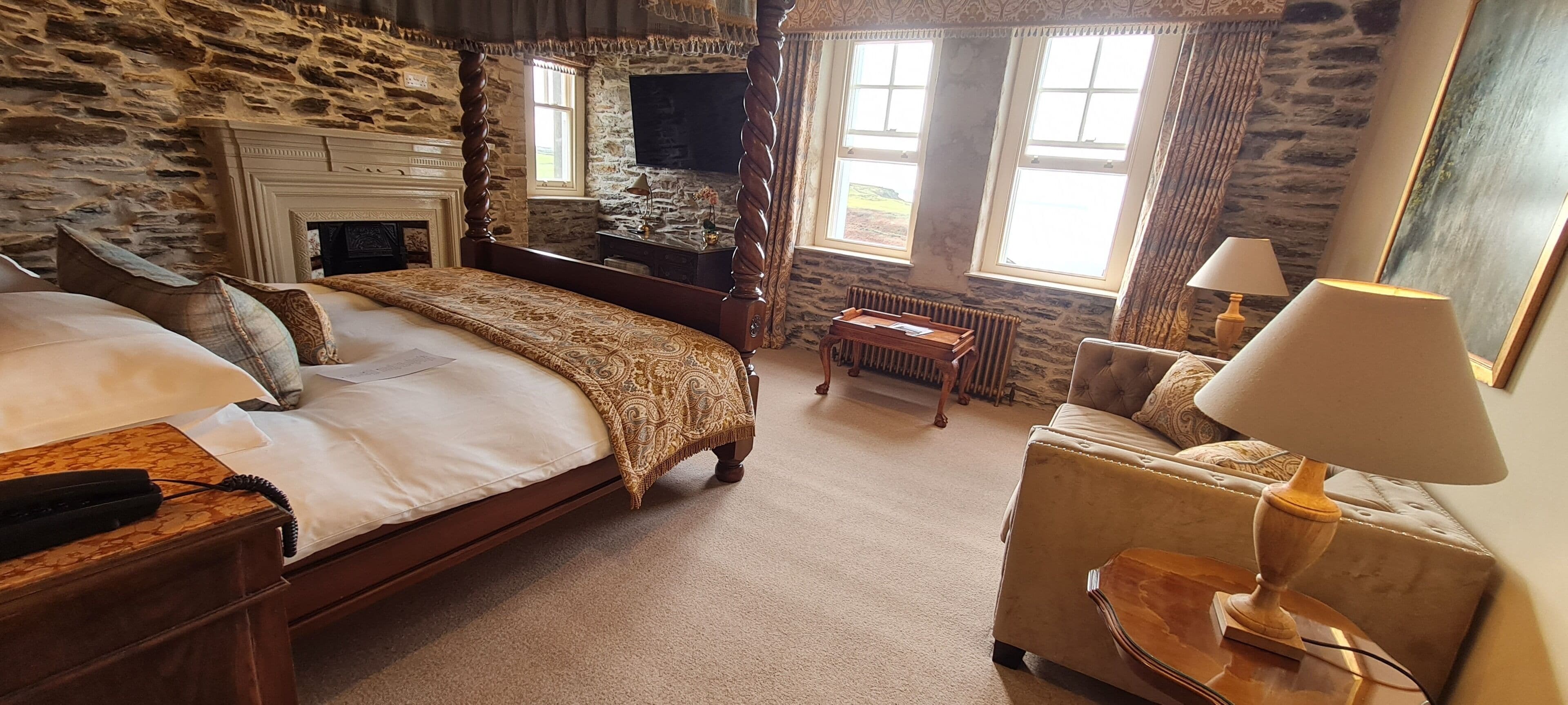 A bedroom with a stone wall, a four-poster bed, a wooden nightstand, a bedside table with a lamp, and a window with curtains.