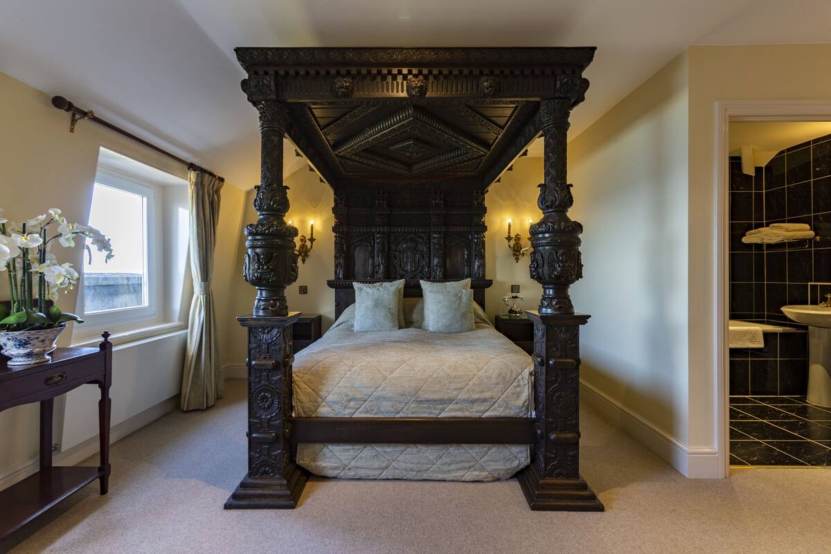 A four-poster bed with a quilted bedspread, a nightstand with a lamp, and a window with curtains.