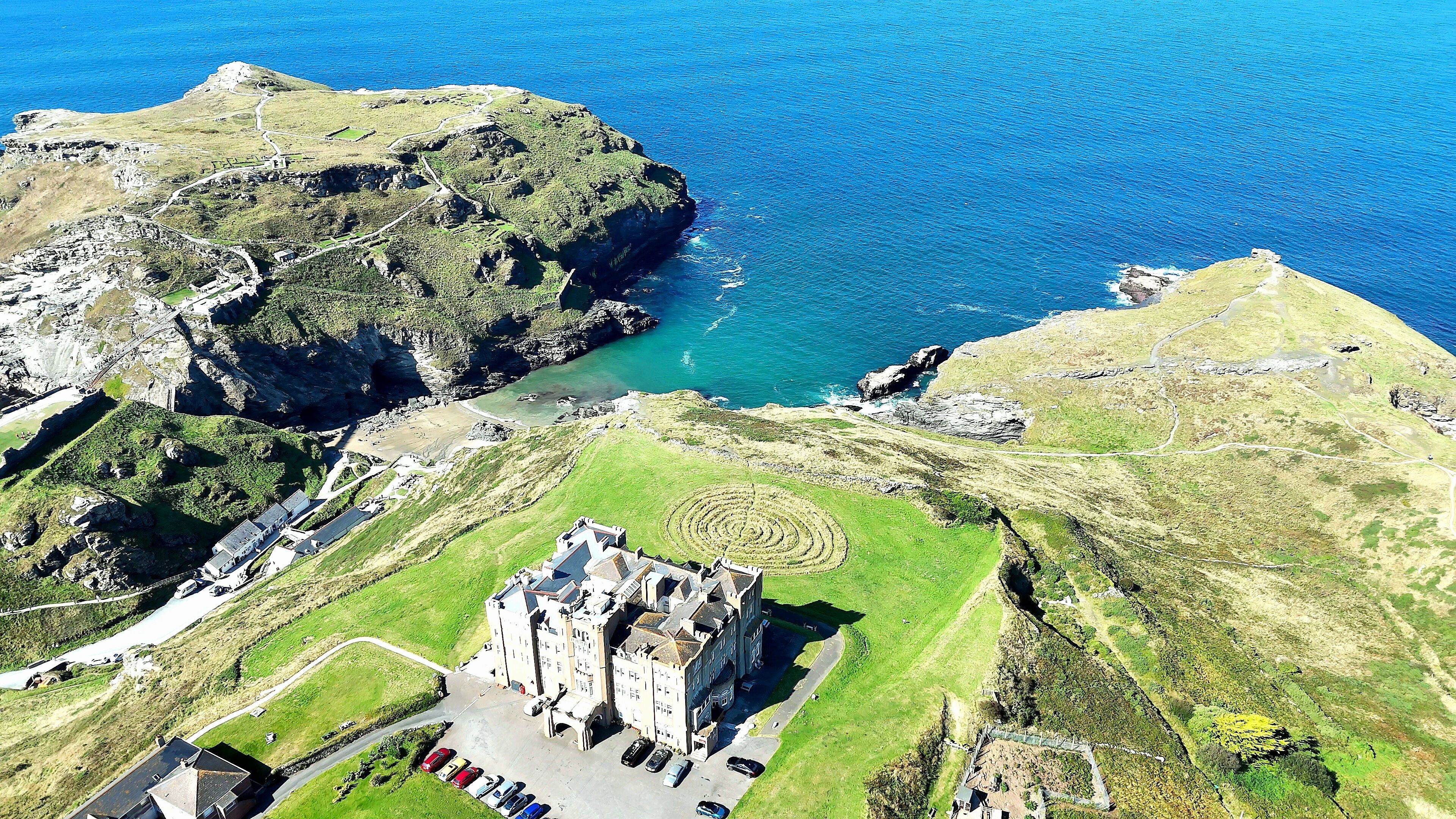 Vista aérea de una zona costera con una estructura similar a un castillo, rodeada de campos verdes y un mar azul cristalino.