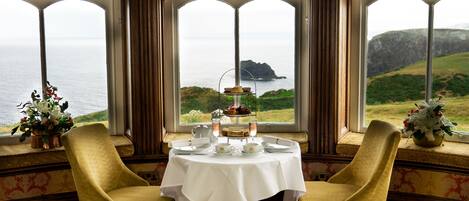 A dining area with a table set for two, overlooking a scenic coastal view through large windows.