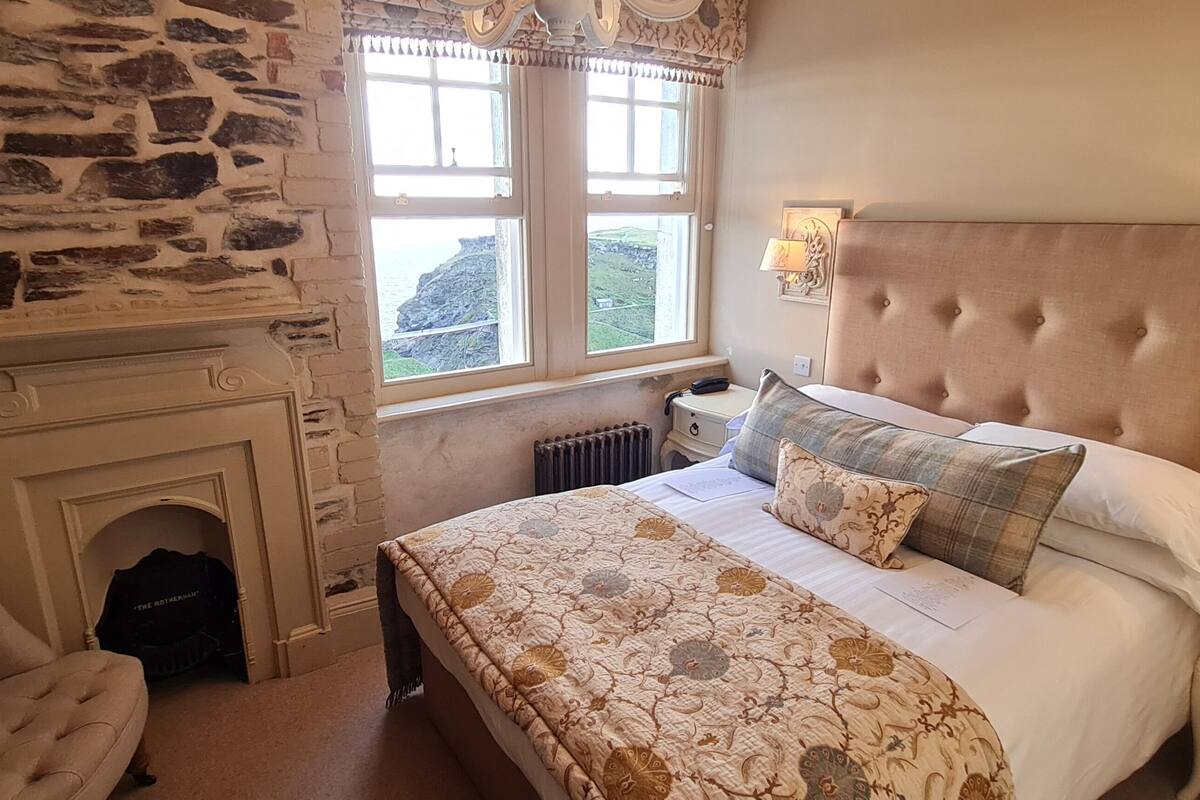 A bedroom with a stone wall, a fireplace, a bed with a tufted headboard, and two wall-mounted lamps.