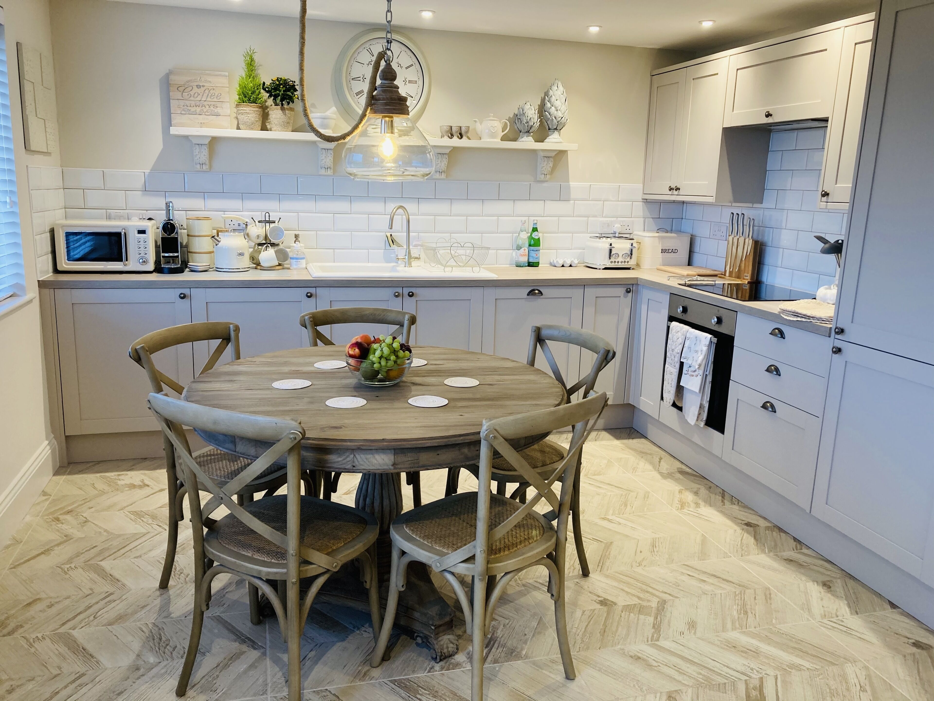 A modern kitchen with white cabinets, a wooden dining table with chairs, a hanging light fixture, and various kitchen appliances.