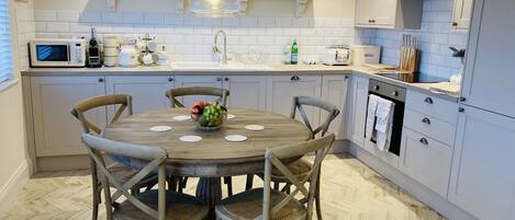 A modern kitchen with white cabinets, a wooden dining table with chairs, a hanging light fixture, and various kitchen appliances.