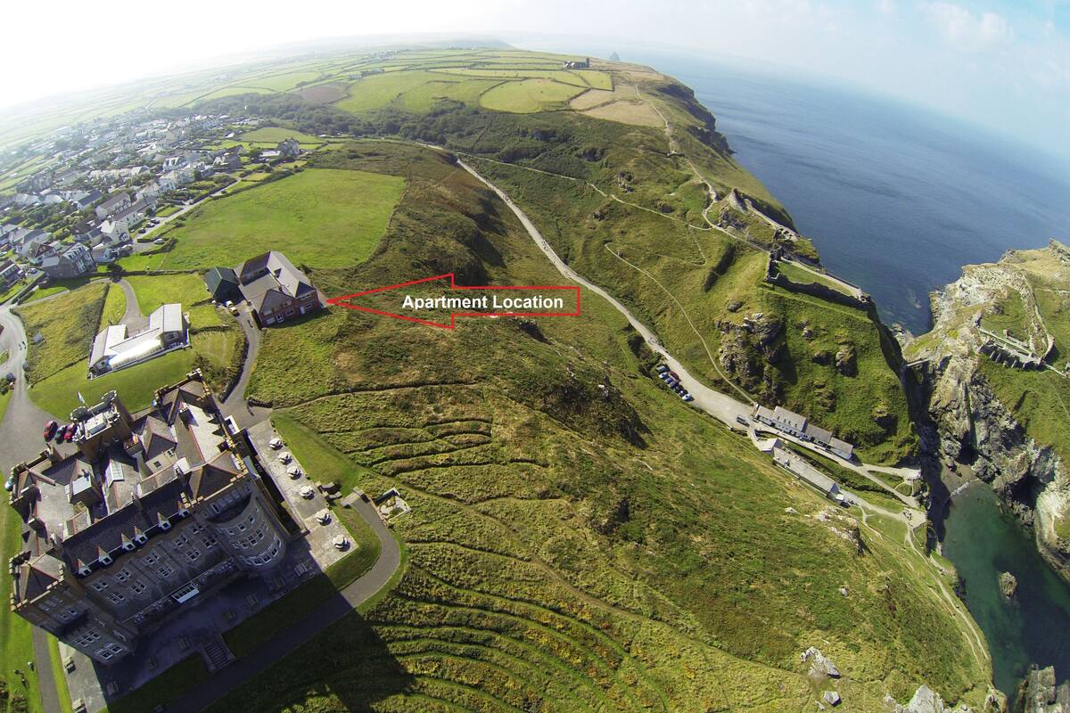 An aerial view of a coastal area with a prominent castle-like building, a winding road, and a marked 'Apartment Location' on a hillside.