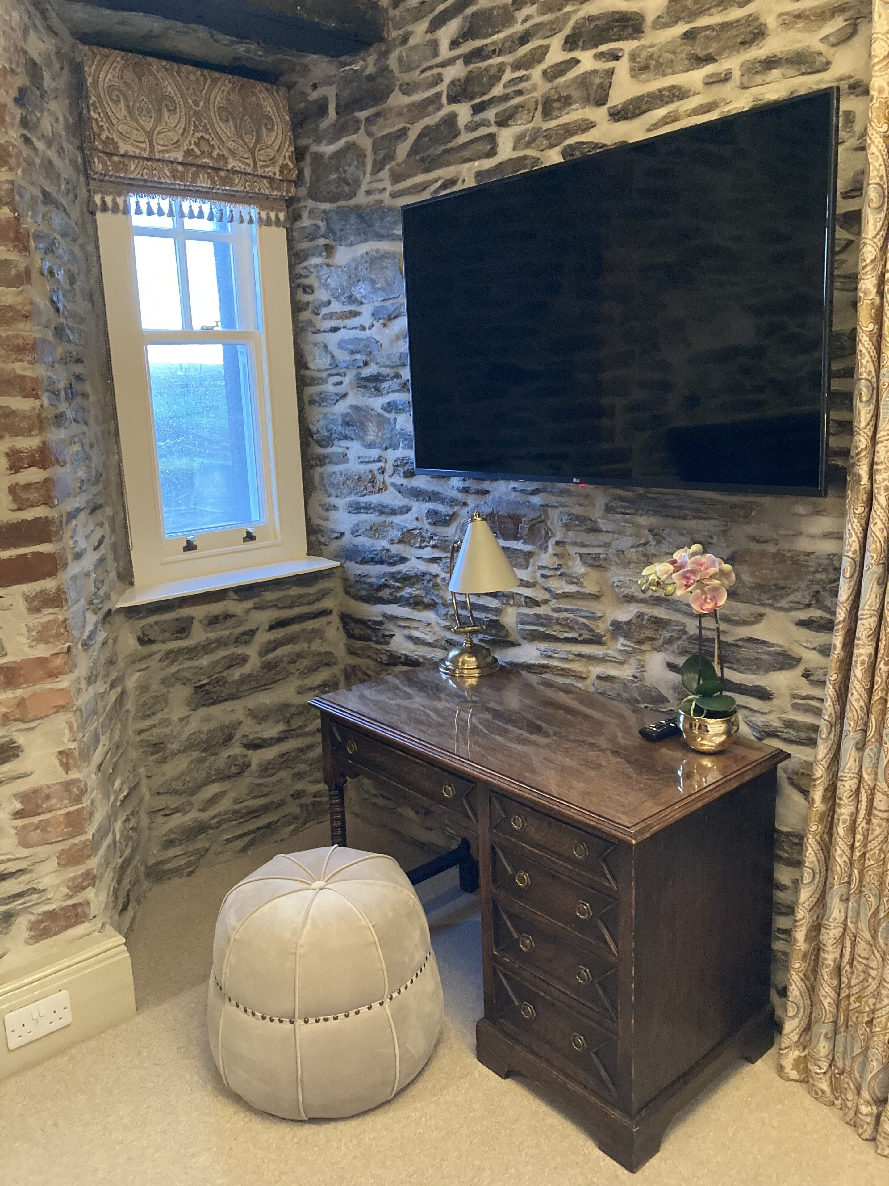 Ein Zimmer mit steinernen Wänden, einem großen Flachbildfernseher an der Wand, einem Holztisch mit Lampe, einem weißen Hocker und einem Fenster mit dekorativem Vorhang.