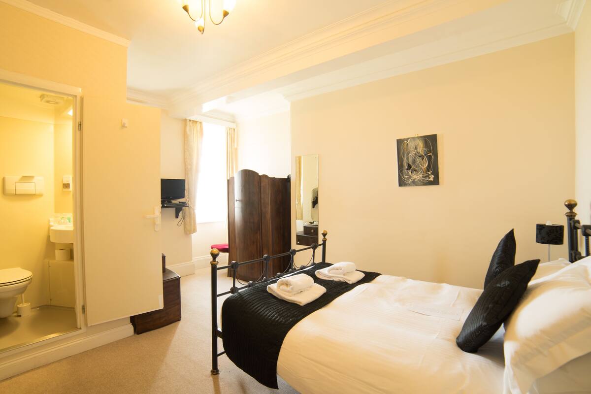 A hotel room with a bed, bedside table, and a mirror. There is a bathroom with a toilet and sink.