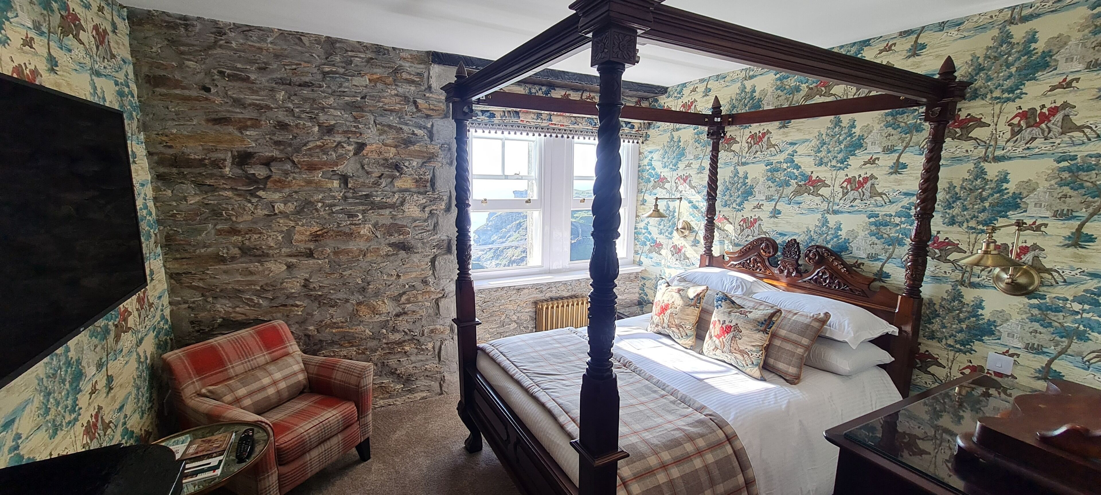 A bedroom with a four-poster bed, a plaid armchair, and a stone wall.