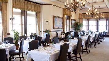 A formal dining room with multiple tables set for service, elegant chandeliers, and large windows with curtains.