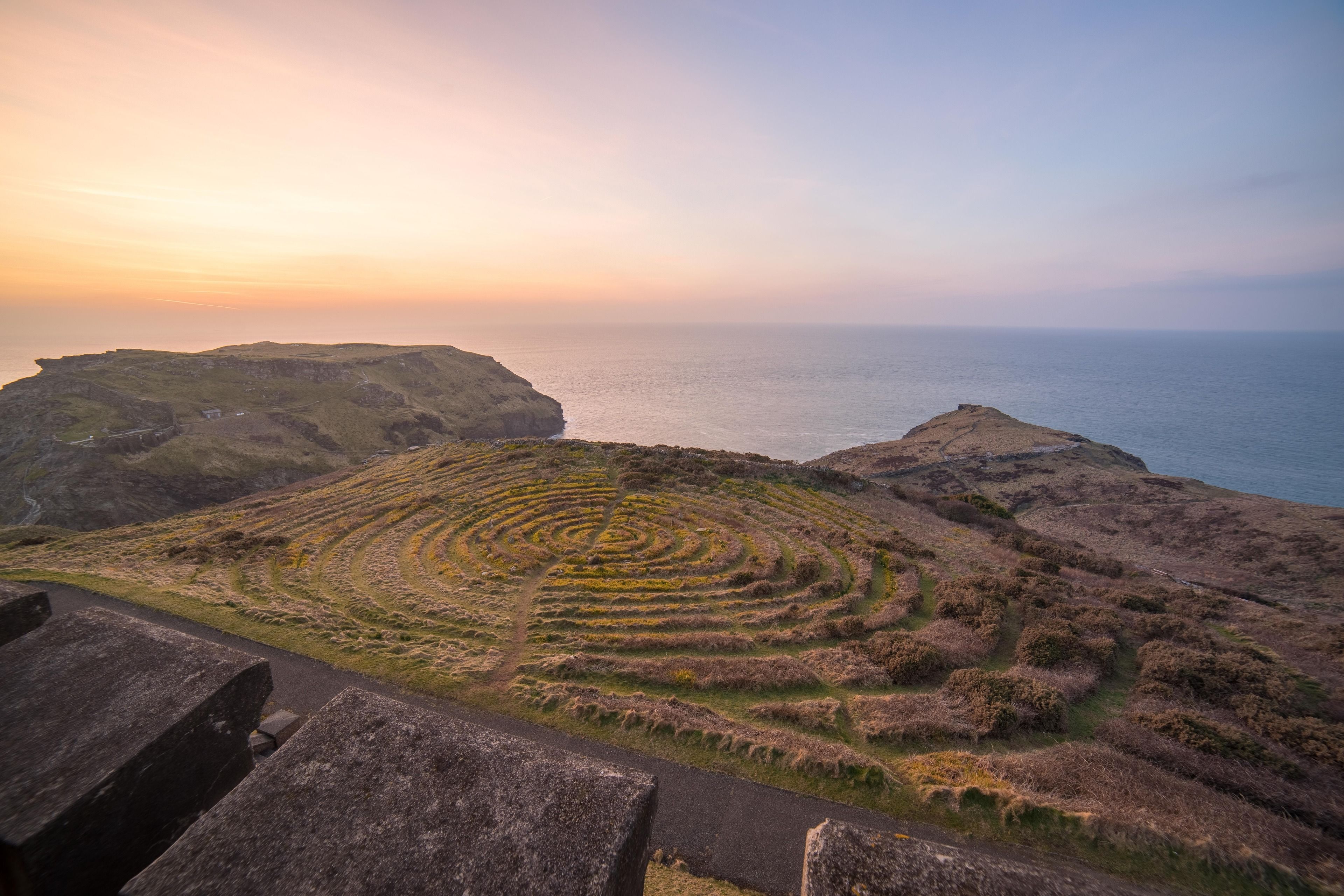 A coastal landscape with terraced fields and a stone pathway leading to a cliff overlooking the sea.