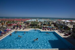 A swimming pool with multiple lanes, surrounded by sun loungers and umbrellas, with a beach and ocean view in the background. - Hotel Sayonara (Cattolica)