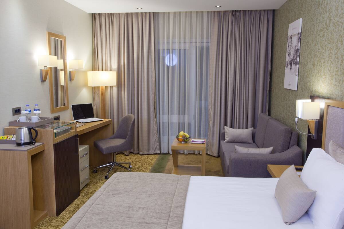 Hotellihuone, jossa on sänky, työpöyt ä tietokoneella, tuoli, pieni pöytä hedelmäkulhon kanssa ja sohva.