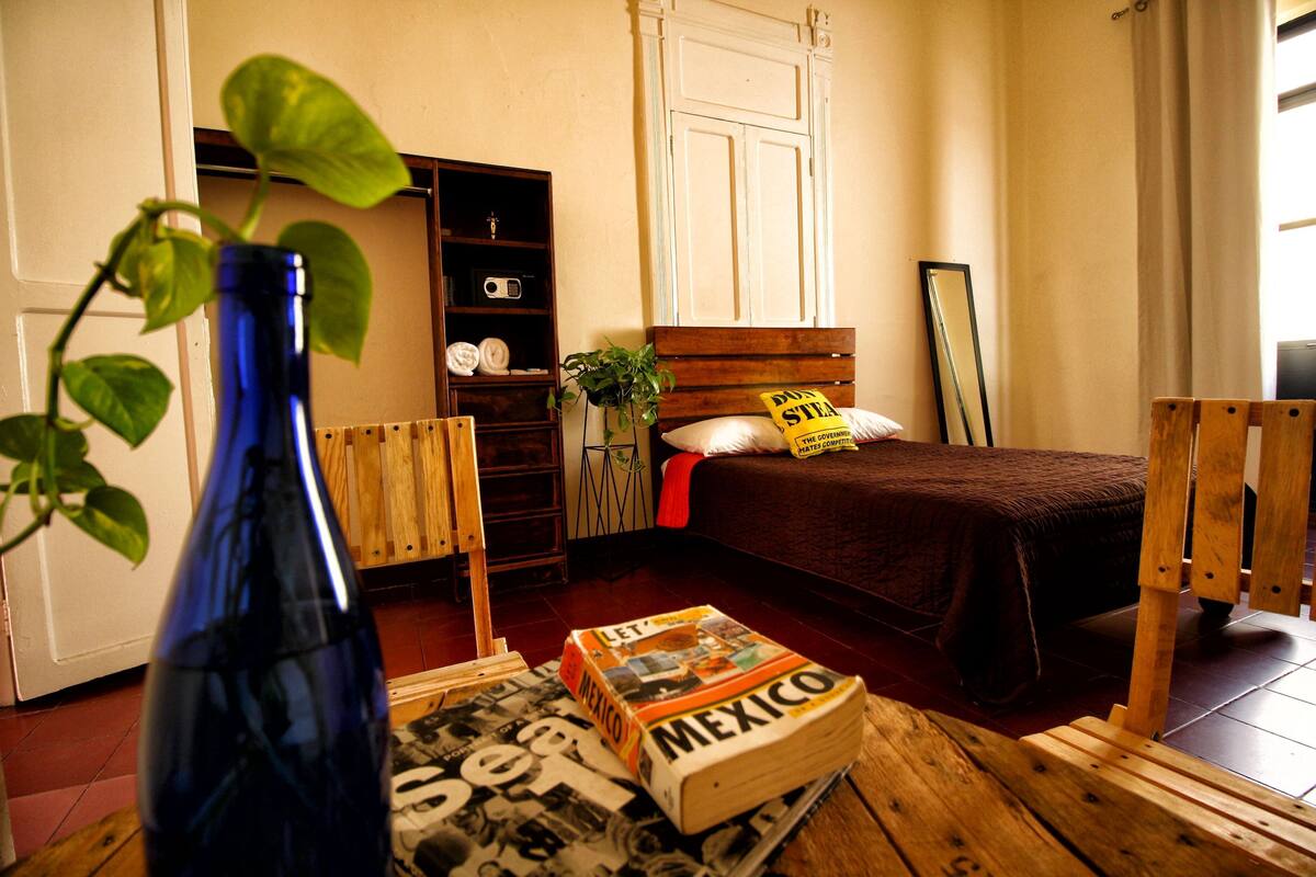 A room with a bed, a wooden chair, a blue vase with a plant, a bookshelf, and a magazine on the floor.