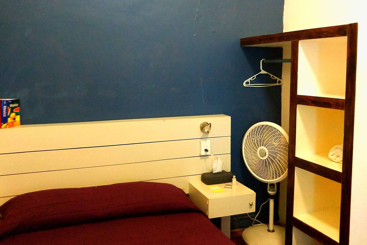 A hotel room with a bed, a bedside table, a fan, and a shelf with a hanger.