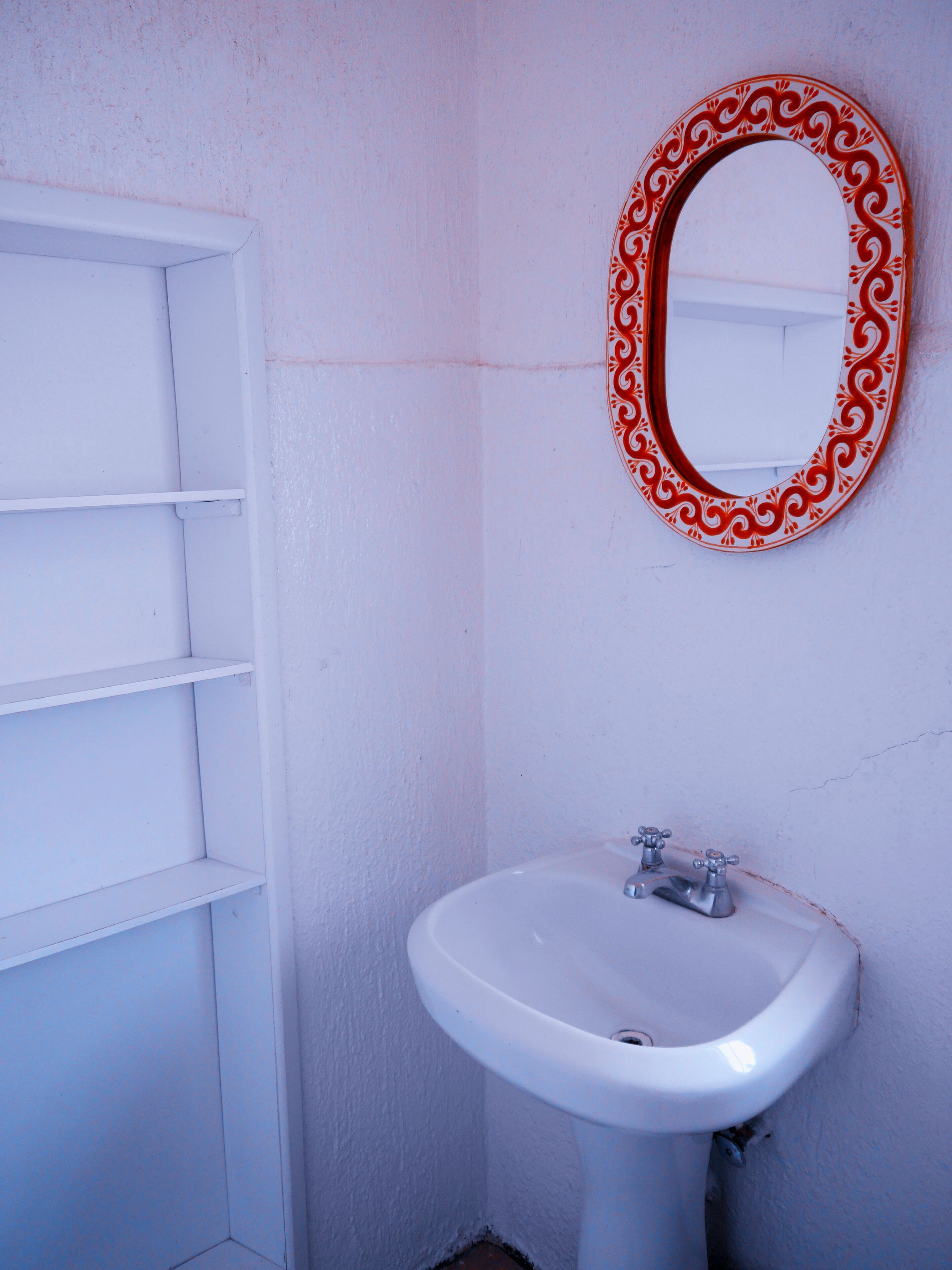 Un baño con un lavamanos blanco, un espejo de marco rojo y una estantería blanca.