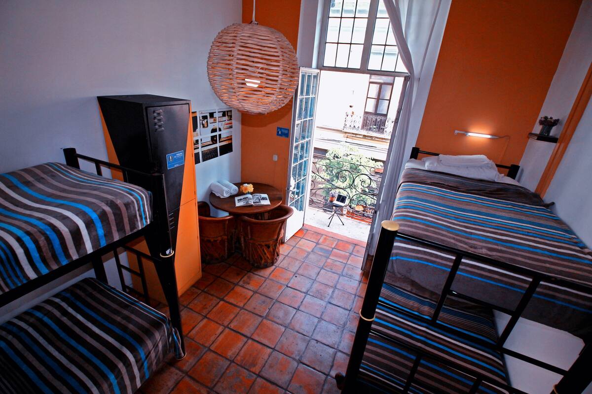 A dormitory room with bunk beds, a desk, a chair, a hanging lamp, and a window with a view of the outside.
