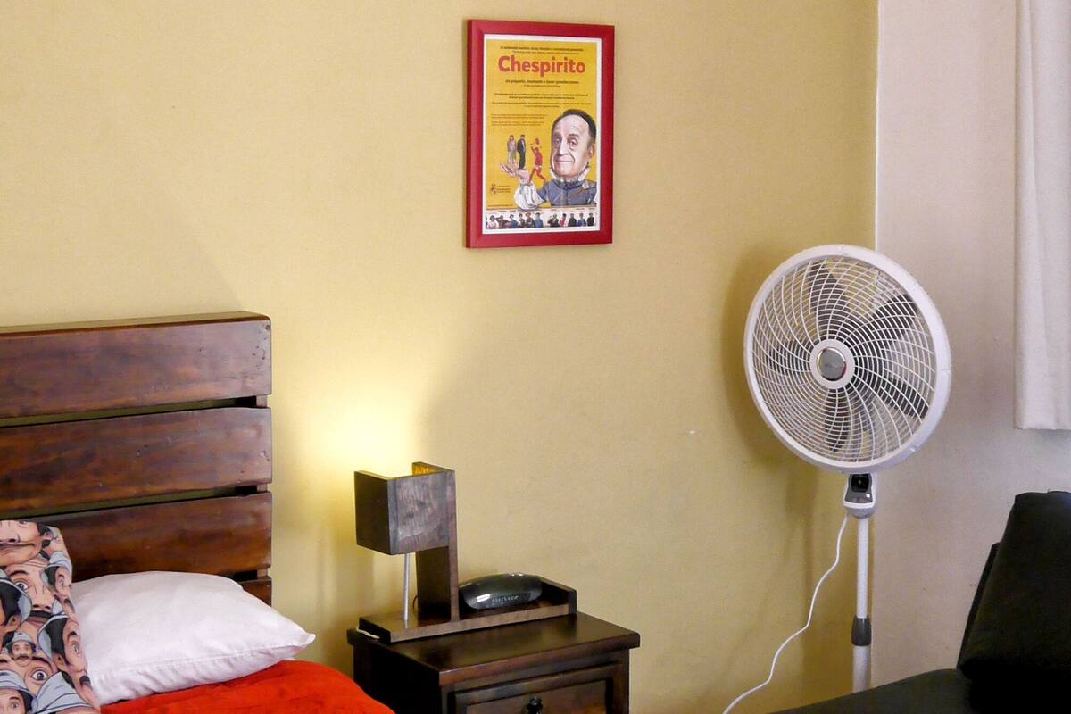A bedroom with a wooden bed, a nightstand with a lamp, a poster on the wall, and a standing fan.