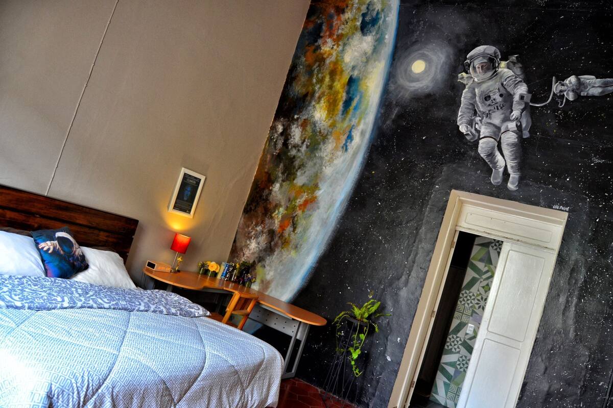 A bedroom with a bed, a nightstand, a lamp, and a wall mural of an astronaut.