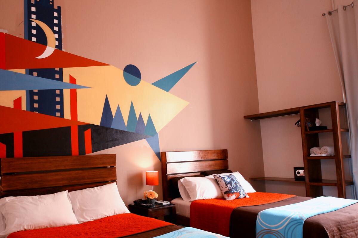 A hotel room with two beds, each with a different colored bedspread. There is a wooden headboard, a nightstand with a lamp, and a wall with a colorful abstract painting.