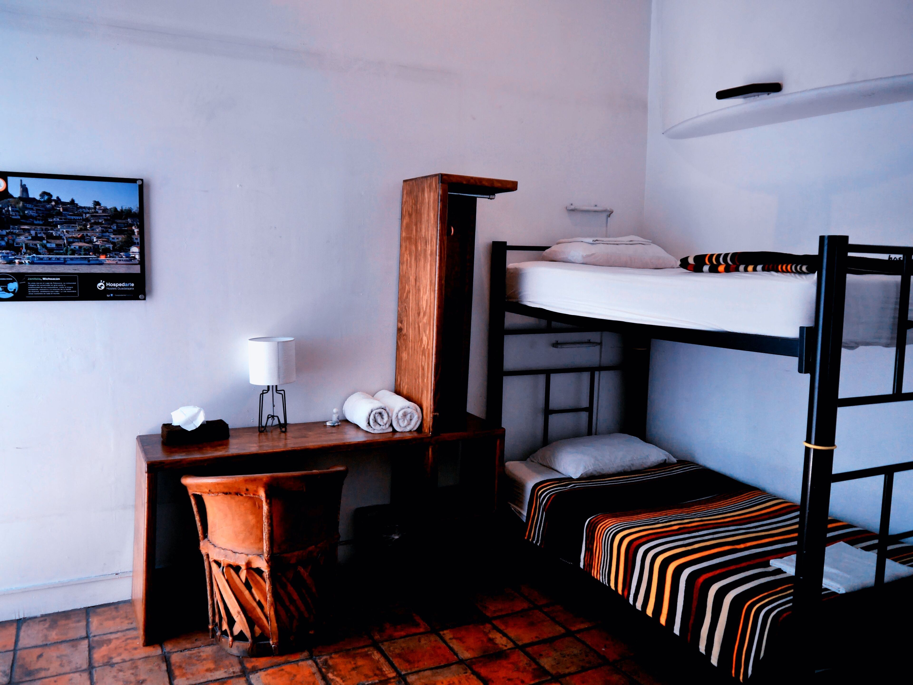 A dormitory room with a bunk bed, a desk, a chair, a lamp, and a framed picture on the wall.