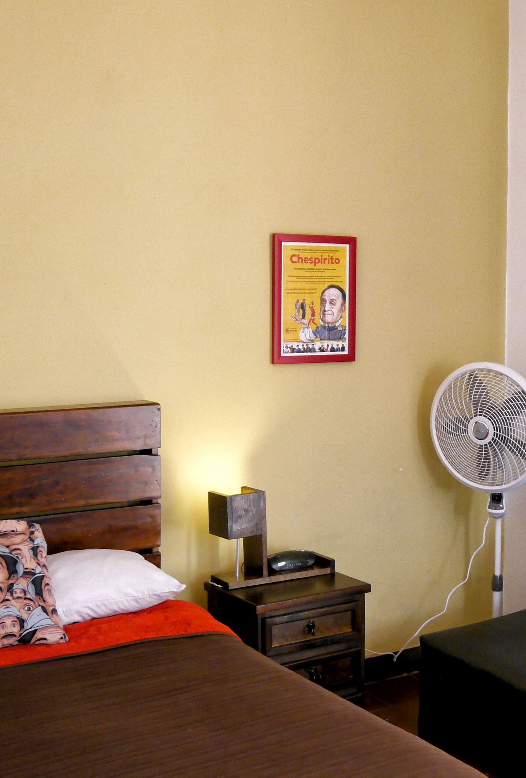 A bedroom with a wooden bed, a white pillow, a red blanket, a small bedside table with a lamp, a poster on the wall, and a standing fan.