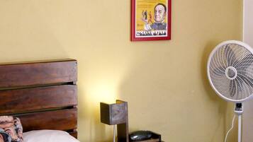 A bedroom with a wooden bed, a white pillow, a red blanket, a small bedside table with a lamp, a poster on the wall, and a standing fan.