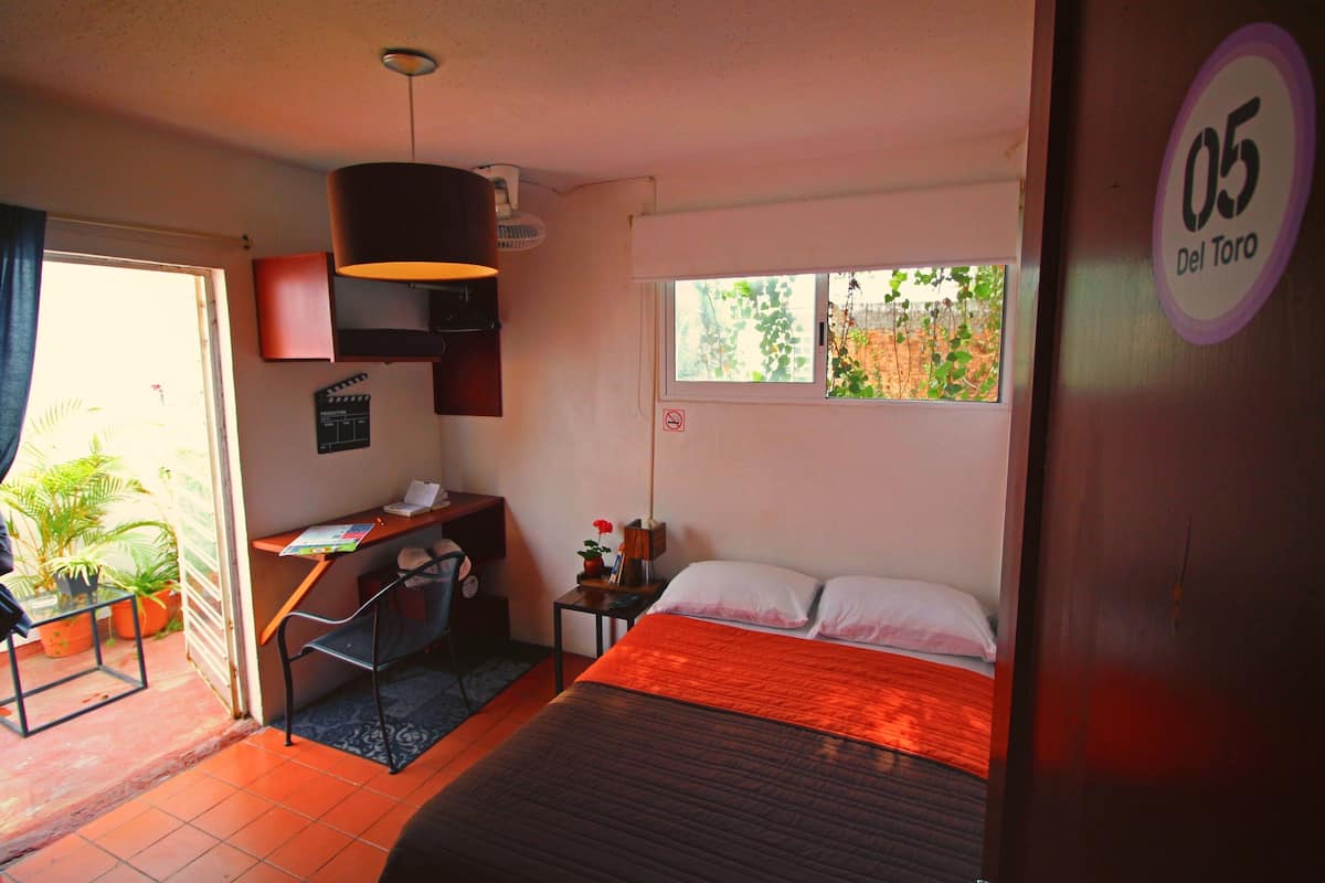 A bedroom with a bed, a desk, a chair, a window with a view of greenery, and a door with a number sign.