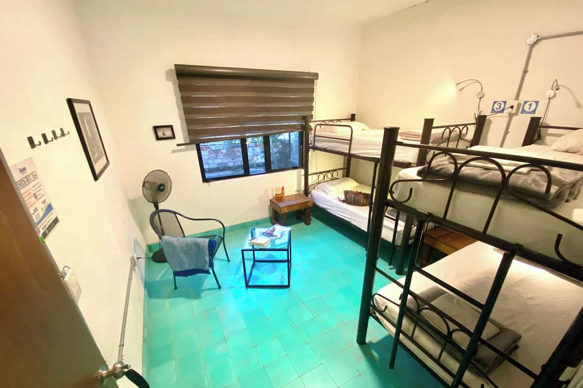 A dormitory room with bunk beds, a small table, a chair, a window with blinds, and a fan.