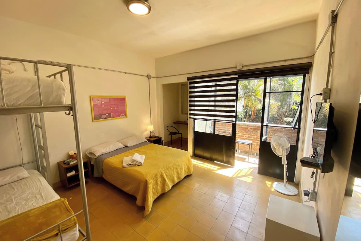 A room with a bunk bed, a single bed with a yellow blanket, a desk with a chair, a standing fan, and a window with blinds.