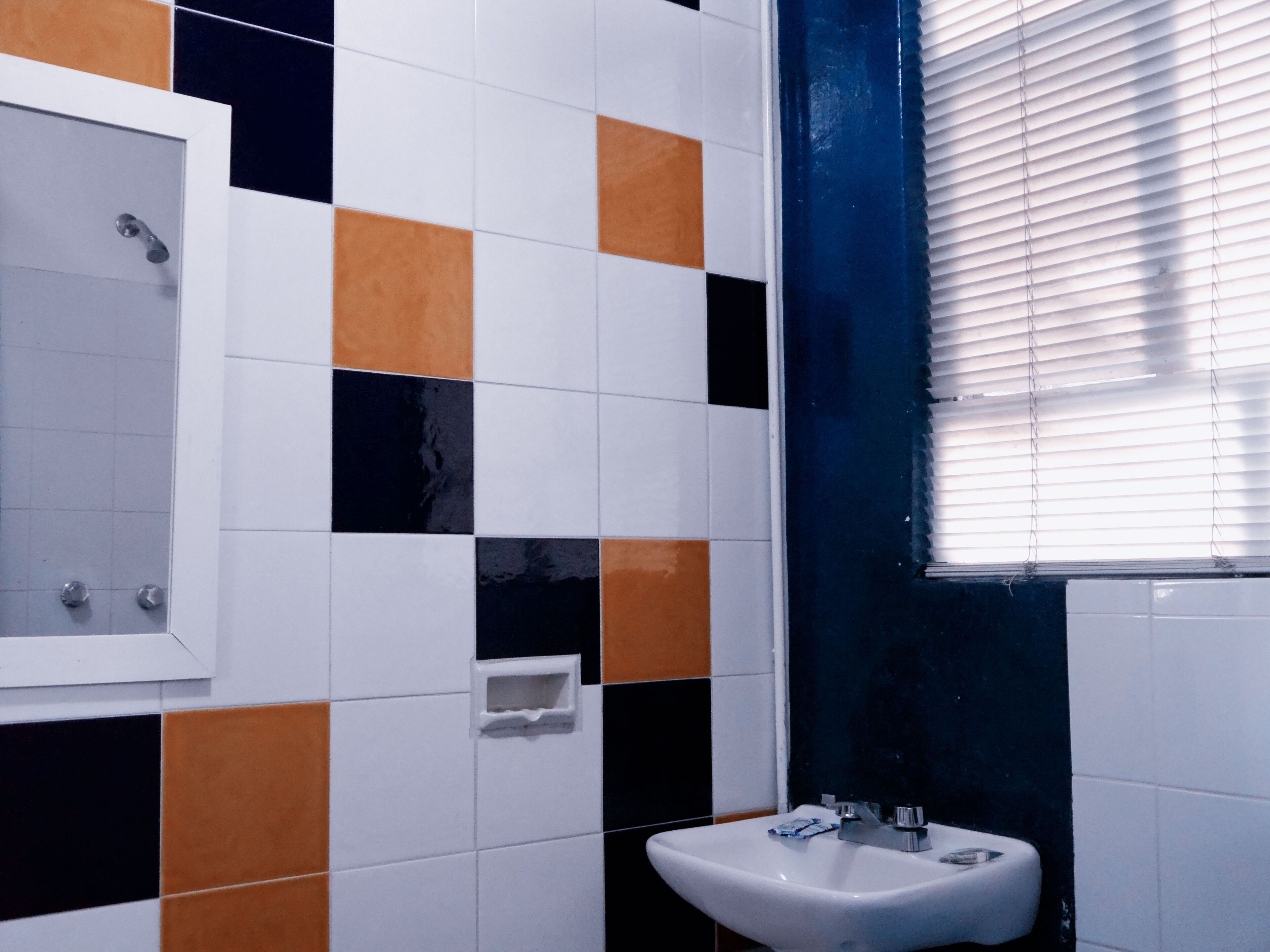 Baño con pared revestida de azulejos con un estampado a cuadros en blanco, negro y naranja. Hay un lavamanos blanco con un espejo encima y una ventana con persianas a la derecha.