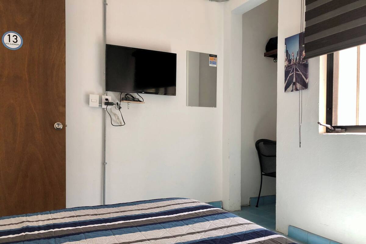 A hotel room with a bed, a television mounted on the wall, a door with a number, and a window with blinds.