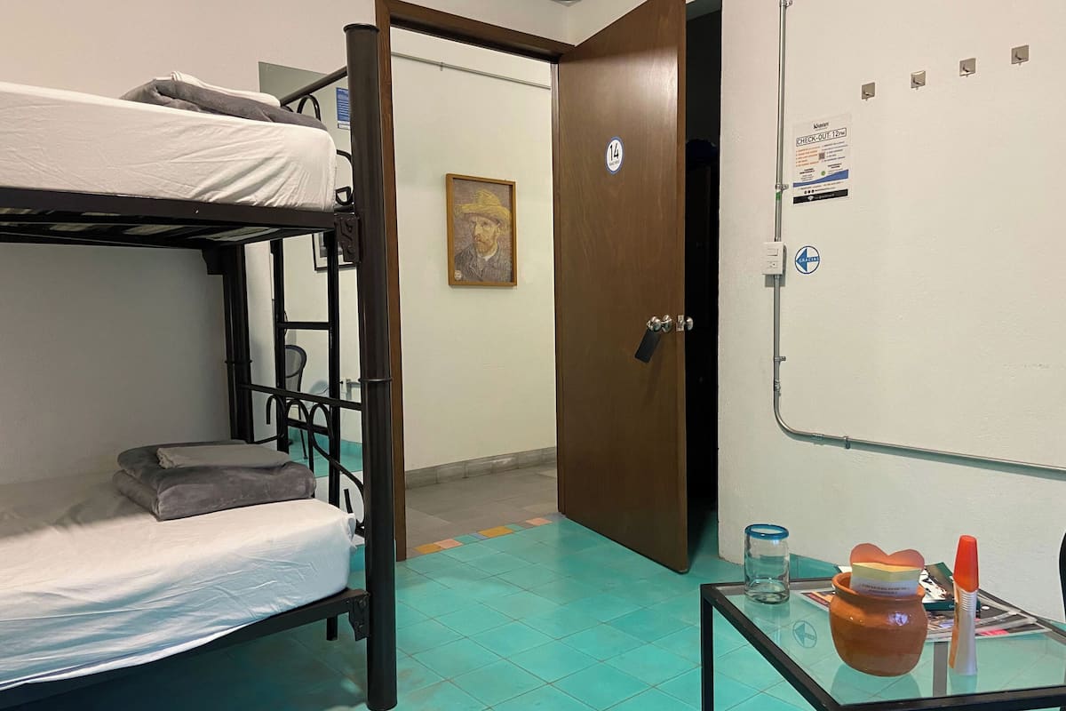 A dormitory room with bunk beds, a wooden door, a check-out sign, and a small table with a vase and a bottle.