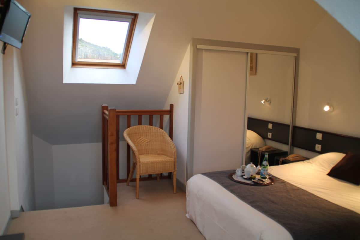A bedroom with a bed, a chair, a small table with a tray, a nightstand, a bedside table, a mirror, and a window with a view of a mountain.