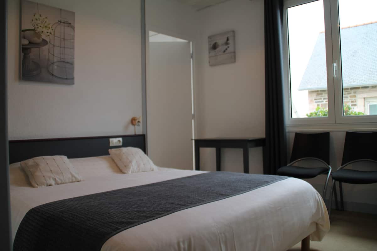 A hotel room with a bed, a small table, two chairs, and a window with a view of a building.