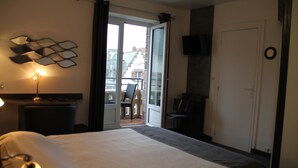 A hotel room with a large bed, a desk with a lamp, a chair, a TV mounted on the wall, and a balcony with a chair and table. - Hotel de la Mer (Pleneuf-Val-Andre)